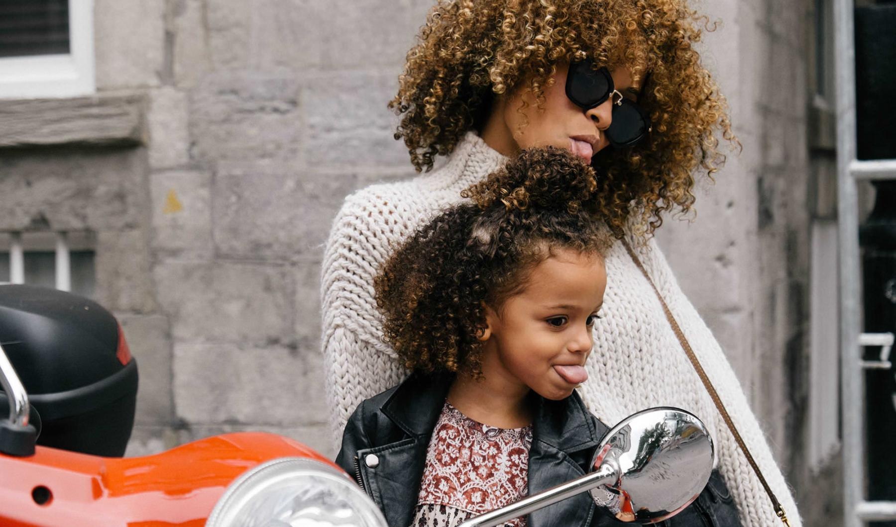 mother and daughter stick their tongues out while looking at themselves in a motorcycle mirror parked in front of a building